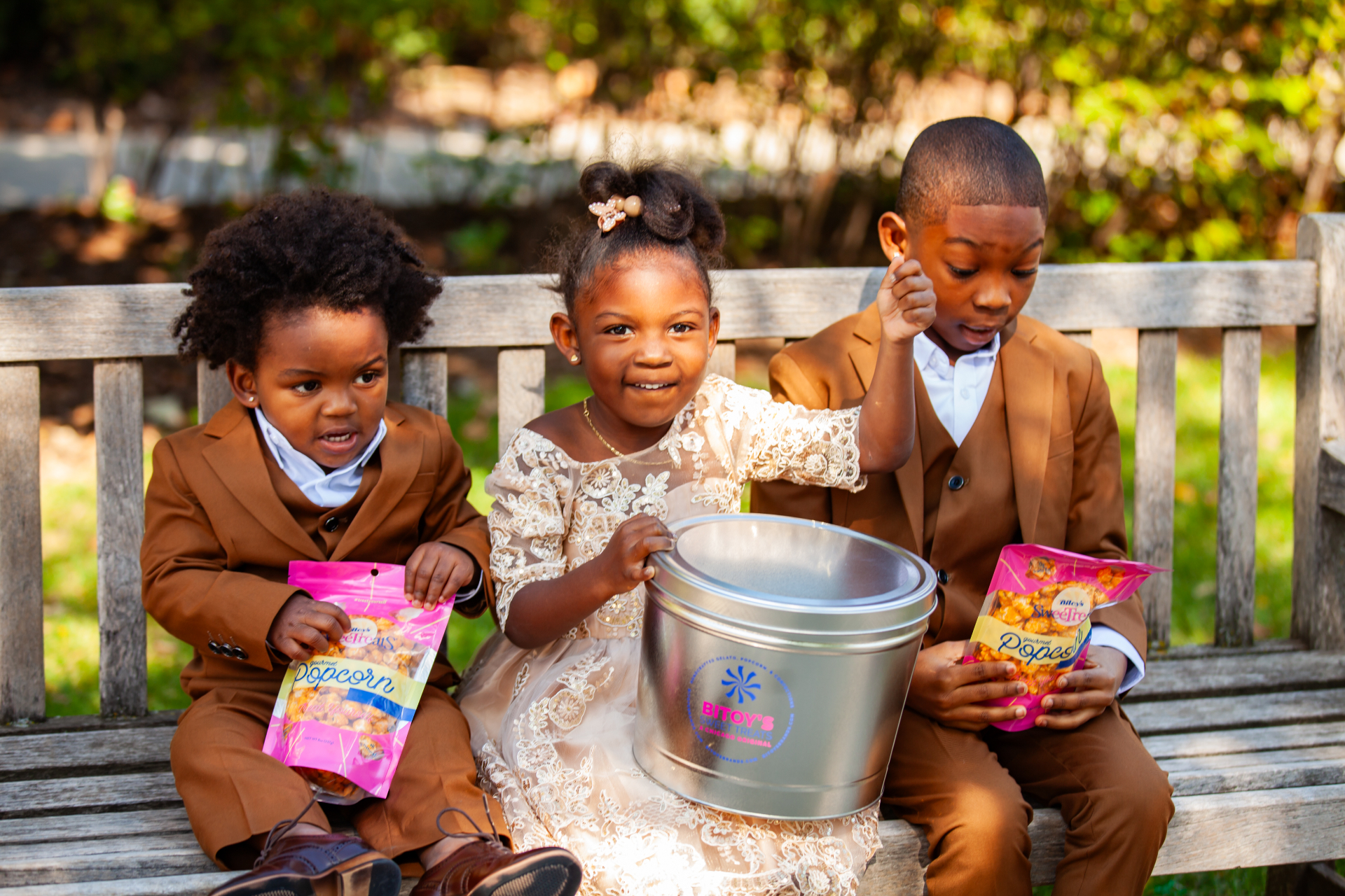 Three children smiling on a park bench with Bitoy's popcorn tins and treat bags